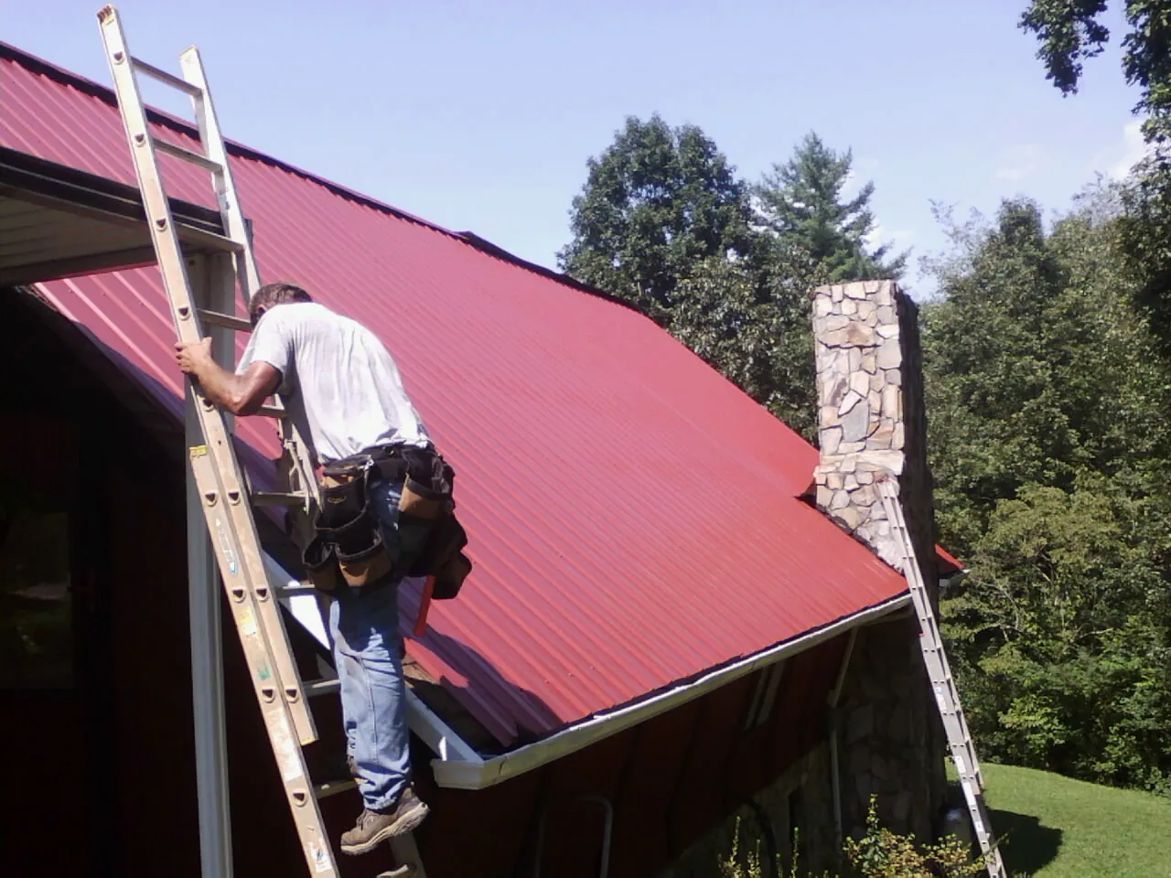Roofer on a ladder beside a red metal roof and stone chimney — Soffit & Fascia Repair by Roofing Brothers in Huntley